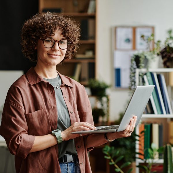 Young successful manager in casualwear standing in front of camera in coworking space and using laptop while analyzing online data