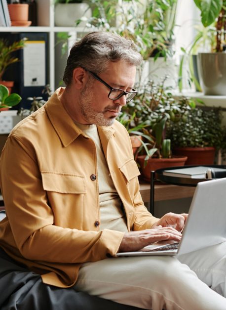 Serious mature businessman in casualwear and eyeglasses with laptop on knees networking in office while sitting on soft bag chair