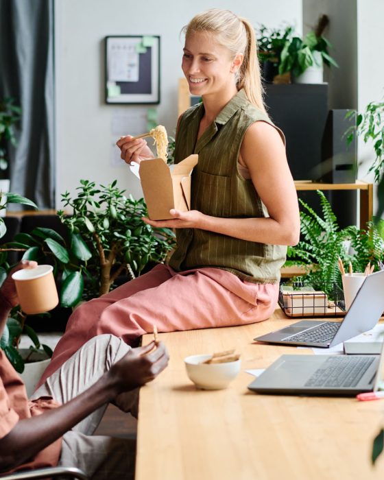 Young smiling female office manager eating wok and talking to colleague Young smiling female office manager eating wok and talking to African American male colleague sitting by workplace among green plants