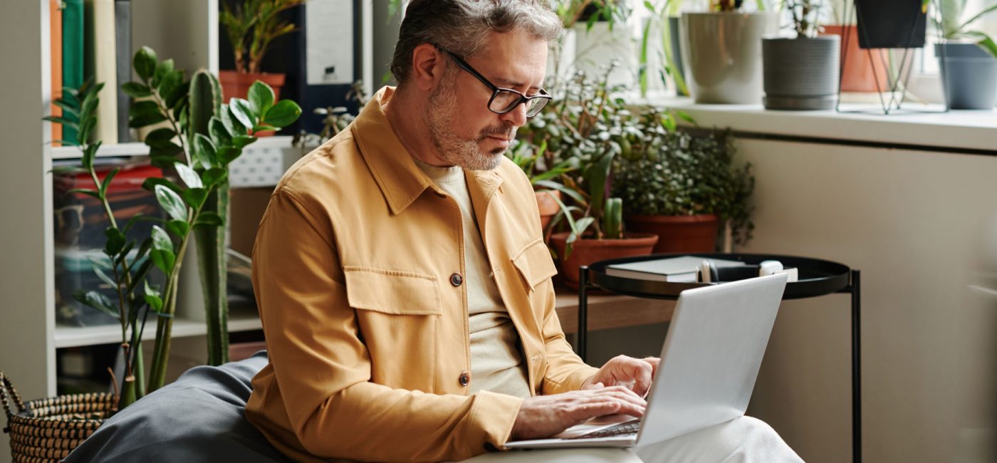 Serious mature businessman in casualwear and eyeglasses with laptop on knees networking in office while sitting on soft bag chair